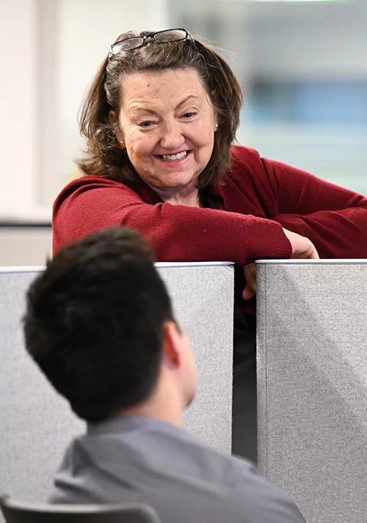 Employee leans over desk to chat with a colleague.